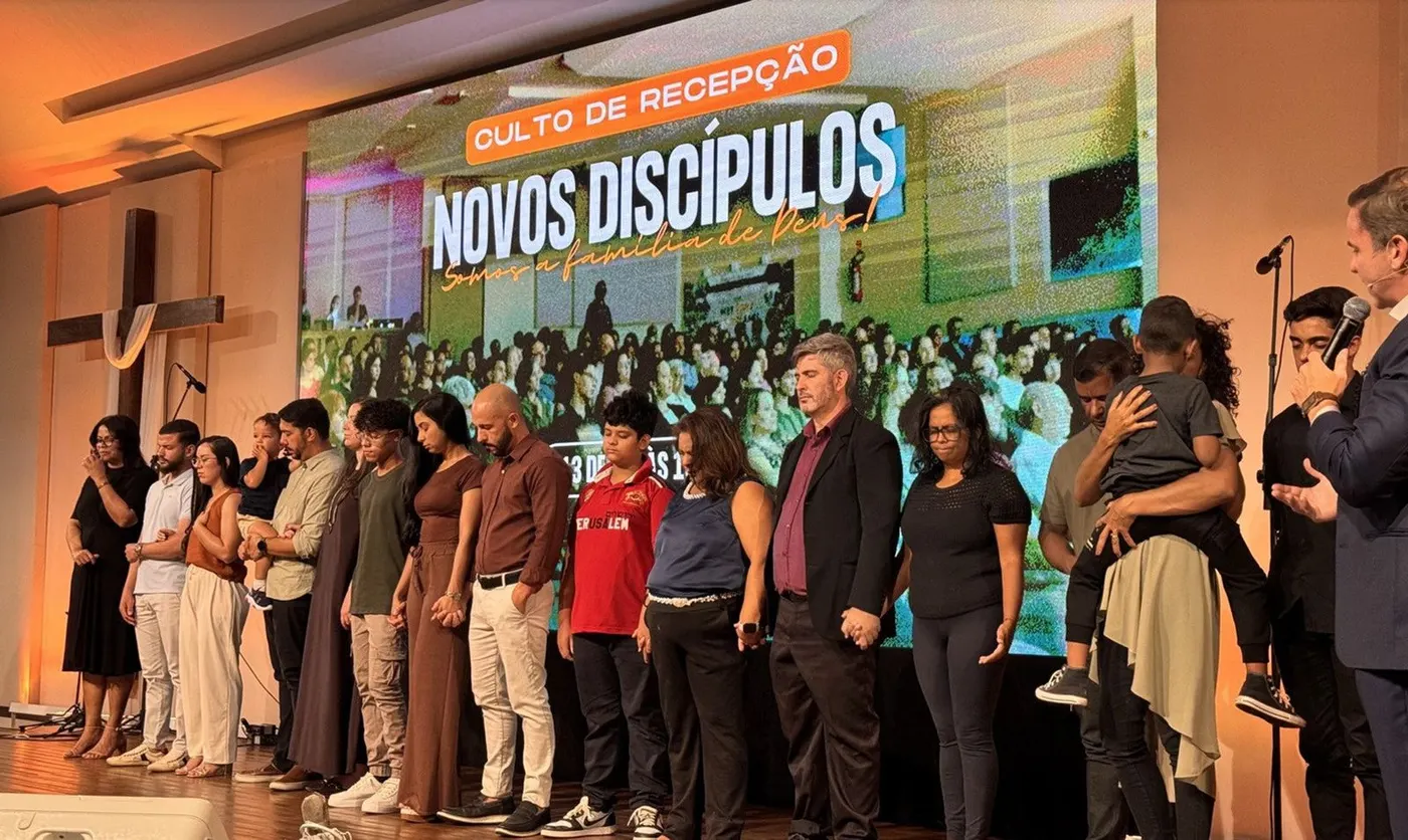 Culto de Recepção: novos discípulos recebidos pela igreja, em oração diante do painel Somos a família de Deus